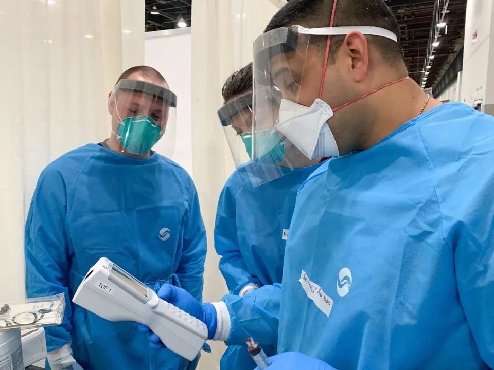 Medical personnel in protective equipment holding an i-STAT 1 handheld blood analyzer.