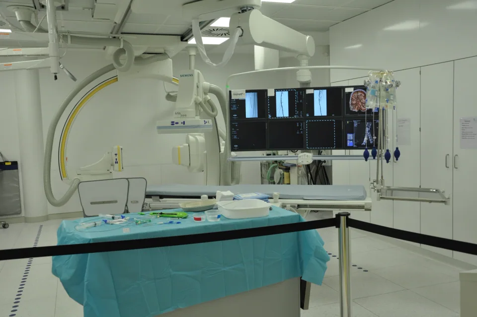 An installed angiography C-arm, radiolucent patient table and overhead monitor in an imaging room.